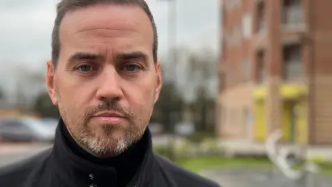 A man in his mid 30s with a light beard looking at the camera wearing a black coat.