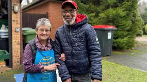 A woman in a blue apron with "Friends of Di's Kitchen" on and a purple shirt links arms with a man in a puffer jacket and red cap. Both are smiling at the camera. 