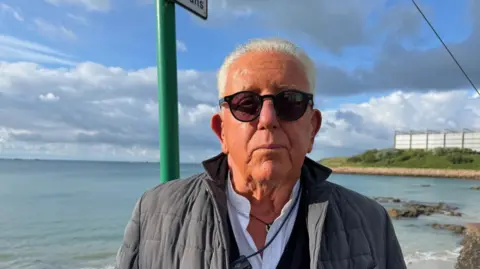 John Morgan pictured in Havre De Pas, he has black sunglasses and a black jacket and a white shirt. 