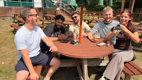 Four men and a woman sat around a circular wooden pub table outdoors with a parasol above them, some with drinks in their hands and two of the men have sunglasses on