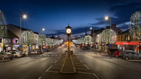 A two way street with trees that are lit up on either side. Shop fronts and cars are along the side. A clock tower is in the centre of the frame, in the middle of the road. 