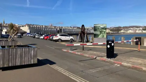 Red and white barriers at the front of an open-air car park. There are terraced buildings in the background lining the Douglas Promenade, with the sea visible on the right. 