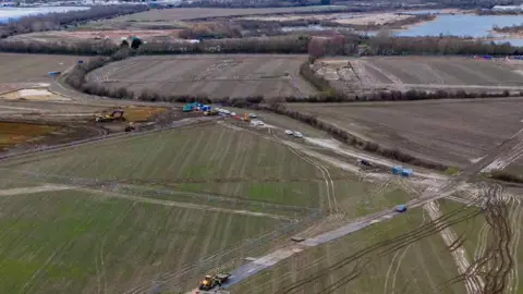 Project Universal A drone shot of the fields at the Universal site, showing construction vehicles on the land including some diggers. There are many track marks in the soil. In the distance are lakes. 