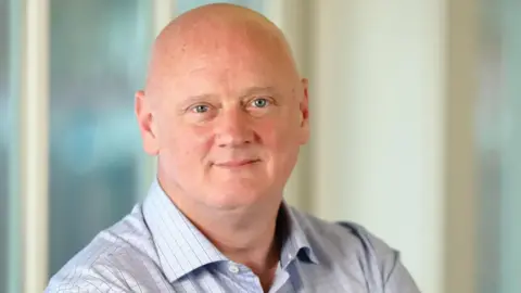 FICO Campbell Scott looking straight at the camera. He is wearing a blue shirt and is standing against a blurred background.