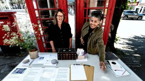 Adam Victor Lisa Wilson and Kim Squirrell standing behind a table next to the two red phone boxes. The table is full of information sheets, clipboards and pens. They are both looking up towards the camera and laughing.