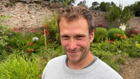 A man with short brown hair smiles at the camera. He is wearing a grey jumper, and is standing outside in front of flowers and a large stone wall. 