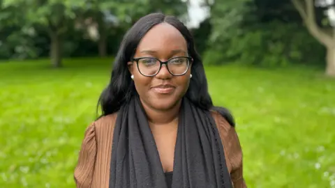 Aaliyah is in a park, standing on the grass in front of some trees and opposite a playground area