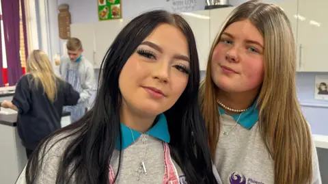 Two young girls wearing grey school jumpers over turquoise polo t-Shirts and standing in a school classroom. They are both looking into the camera. One has dark hair past her shoulders and the other mousy blonde.