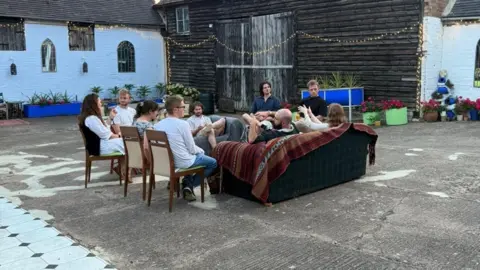 Nine people sat on wooden chairs and a sofa in the middle of a courtyard. 