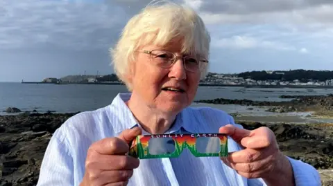BBC Dr Jean Dean holding a pair of the brightly coloured solar eclipse glasses. She has short grey/blonde hair and is wearing a blue shirt. She also has slim framed silver glasses. She is stood in front of a coastal view of Guernsey. 