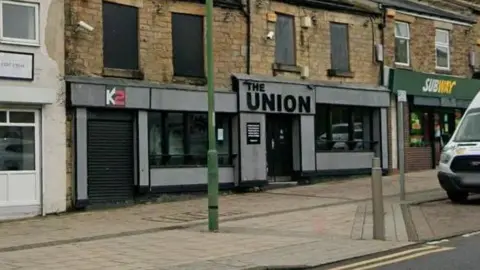 A Google Street View of K2 and The Union in Consett. The two units are next to each other on the lower levels of terraced buildings, flanked by other shops and commercial units, including Subway. Both have grey panelling around the windows and doors where the shop signs are displayed.
