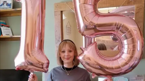 Family handout A teenage girl with shoulder length blonde hair wearing a silver necklace and grey jumper smiling into the camera. She is stood in a room of the house holding up two pink balloons, one says 1 and the other says 5.