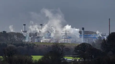 The Sellafield site pictured from a distance. Plumes of white smoke hang in the air and a number of rectangular buildings can be seen along with five chimney stacks. Low circular storage containers sit in front of the buildings.