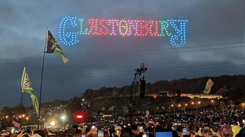 A colourful light show in the sky that spells out the word 'Glastonbury' while crowds of people watch below, taking out their phones to film it