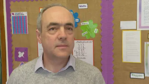 Head teacher Tim Hughes, who is wearing a grey jumper with a striped white shirt underneath. He is a white man with dark eyebrows and grey hair. He is looking toward the camera. He is stood in front of a noticeboard which is brown and purple.