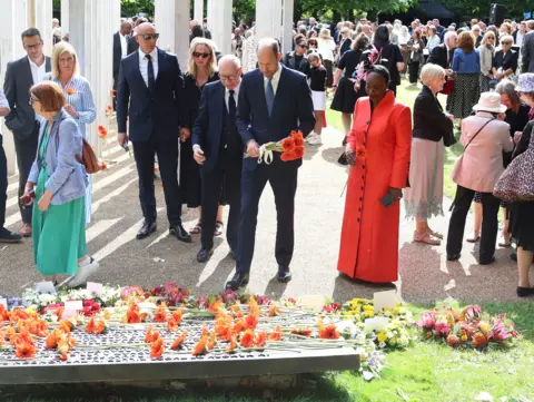 Prince William holds a bouquet of red flowers at a memorial service in Hyde Park. There is a group of people behind him all talking to each other. He is wearing a navy suit. Stood on his left side is Thelma Stober, a survivor of the 7/7 attack. She is wearing a red dress.