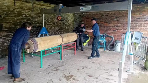 Sheffield Museums Three people work inside a historic workshop with exposed stone and brick walls, handling a large wooden beam mounted horizontally on support stands. An overhead lifting gantry and hoist are positioned above the beam, while industrial equipment and machinery are visible around the room. The workshop has a stone-tiled floor and appears to be undergoing restoration or conservation work on the timber structure.