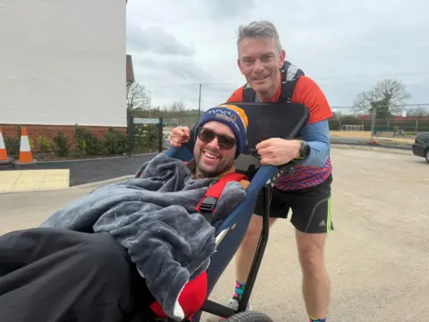 Two white men looking and smiling at the camera, one is in a wheelchair the other stands holding him.
