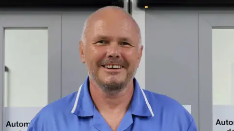 Paul Johnson has very short cropped and receding grey hair, and a beard. He is wearing a blue nurses uniform and is stood in front of double doors inside a hospital ward.