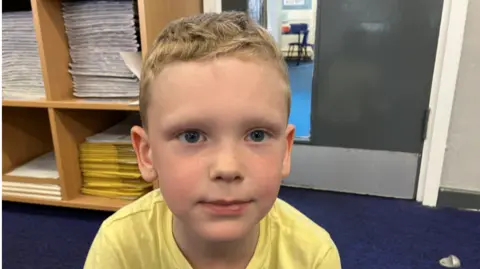 Theo is wearing a yellow t'shirt and has blonde/brown hair. He is sat on the floor of his classroom and almost smiling at the camera.
