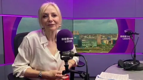 A woman with blonde hair and wearing a cream blouse sitting in a radio studio with a purple backdrop