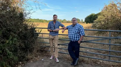 Two men, both wearing patterned blue and white shirts, trousers and trainers, stand against a metal gate with a field behind them and trees surrounding the area.