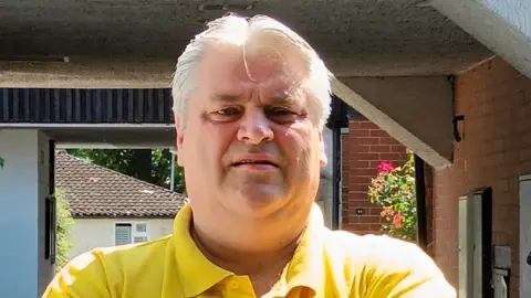 A man with very blond hair and wearing a yellow polo top looking slightly down into the camera. There is a porchway in the background and a house beyond that.