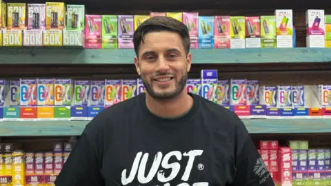 A man with dark brown hair stood in front of shelves stacked with vapes.