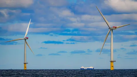 A view of two offshore wind turbines in the sea. A ship can be seen sailing past in the distance. Clouds dot the sky.