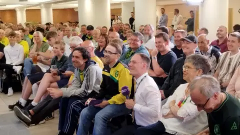Henry Miller/BBC Seated people smile as they listen to speakers addressing their questions in a room.