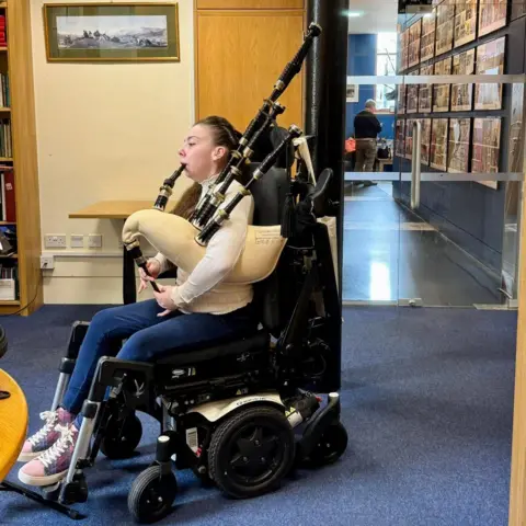 Katie Robertson A woman in an electric wheelchair playing bagpipes in a corridor with blue floors.