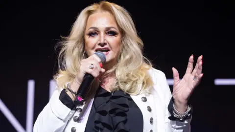Bonnie Tyler on stage holding a microphone. She has long blonde hair, and is wearing a black blouse and a white jacket