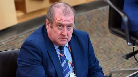 Getty Images Graeme Dey sitting in the Scottish Parliament. He is wearing a dark blue suit, light blue shirt and a blue striped tie.