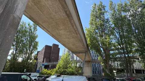 A tall concrete bridge, with concrete columns, spans a car park and connects to further concrete walkways. There is a road sign, tall trees and tall buildings in the background.