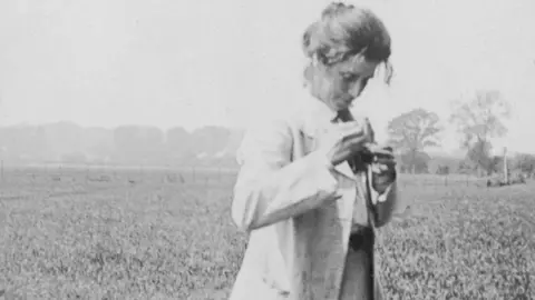BPEOSA collection Margaret Stansfeld, in a field, looking down putting a daisy in her lapel. She has her hair tied up, and is wearing a long coat. 