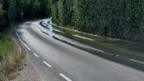 Malcolm Owers A road where the right-hand side is covered in a thin layer of water from a burst water pipe. The road is lined with thick bushes and trees.