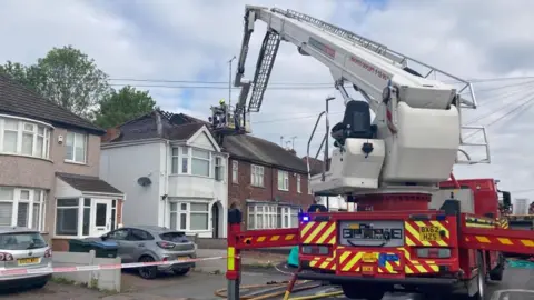 A fire engine and hydraulic platform on a residential street. A firefighter is on the platform next to a fire damaged roof.