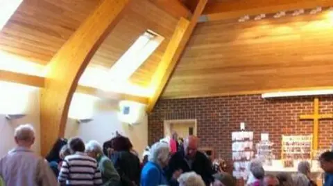 The interior of the chapel at the Homestead with a large cross at one end on a brick wall. There are several people sitting on red wooden-framed chairs, or standing between them. There are large lights on a curved wall to the left.