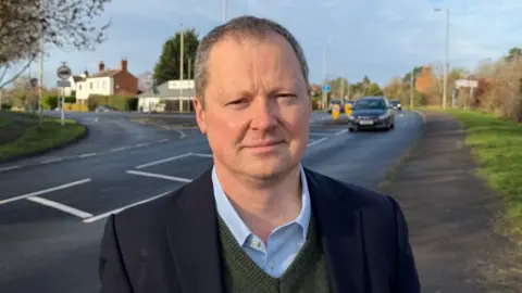 A head and shoulders image of a man in a jacket standing by a road
