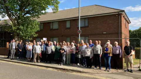 LDRS Group of 40 odd people standing outside a brick building looking at the camera. They are all dressed in various clothes. It is a sunny day.