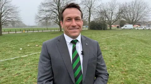 Richard Lewis wearing a grey suit with a black and green striped tie. He is standing on a grass rugby pitch, and has a cut above his left eye sustained in a recent rugby match.