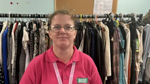 Jane Platt in a bright pink polo shirt. It says Big Venture Centre. Her lanyard says the same. She has glasses and is smiling. There are lots of clothes on a big rack behind her. 