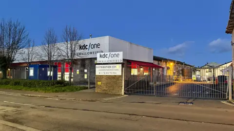 The outside of the Swallowfield site, which is a low square building behind a large fence. The KDC/One logo is visible on the building and on the gate.