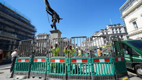 PA Media The statue is seen behind grey and green barriers, which are being adjusted by several men wearing hi-viz jackets.
