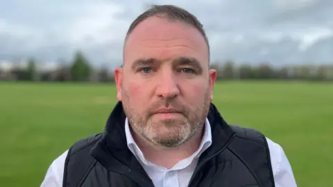 Sam Read/BBC Finbarr Fitzpatrick stands in a park with a grass field on a grey day. He has short dark hair, stubble, and wears a white shirt with a black gilet jacket over the top. 