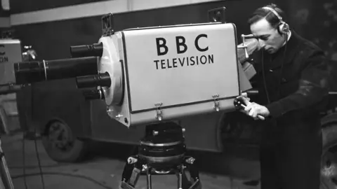 A black-and-white shot of a large, old-fashioned television camera, pointing to the left of frame. It has 'BBC Television' written on the side, and has a turret lens set-up, with which to get different focuses a different lens must be physically rotated into place. On the right of frame, a male operator is holding the camera and looking through its viewfinder.