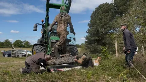 Jamie Niblock/BBC A bronze statue of a man is transported by a green tractor at the edge of a field. Two men on their knees handle the bottom of the statue. Another man looks on as they work. Cars are parked in a field behind a gate nearby.