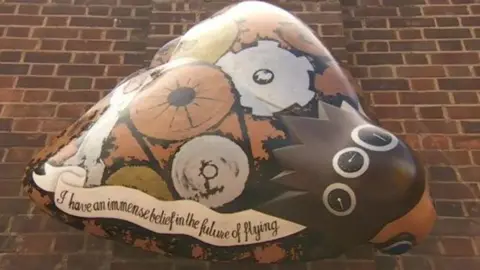 A large moth-shaped sculpture attached to a brick wall. It is painted to celebrate the engineering work behind a plane, with cockpit gauges, wheels, cogs and spanners. The colours are predominantly shades of brown, orange and white. A painted slogan reads: "I have an immense belief in the future of flying".