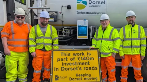 Dorset councillor Jon Andrews (second from right) with three workers from the gully tank and crew. They all wear helmets and high vis uniforms. They are standing around a yellow sign that reads This work is part of £140m investment in Dorset's roads.
