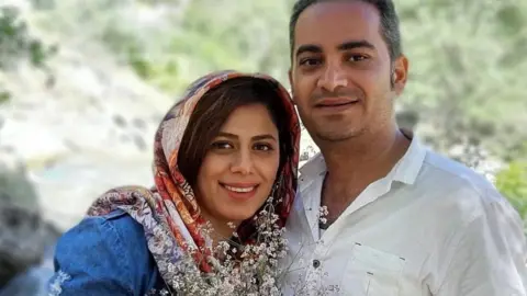 Mansoureh Heydari and her husband, Behrouz Mansouri, smiling and embracing as they look at the camera. Mansoureh wears a headscarf and holds a bouquet of flowers, Behrouz wears a white shirt.